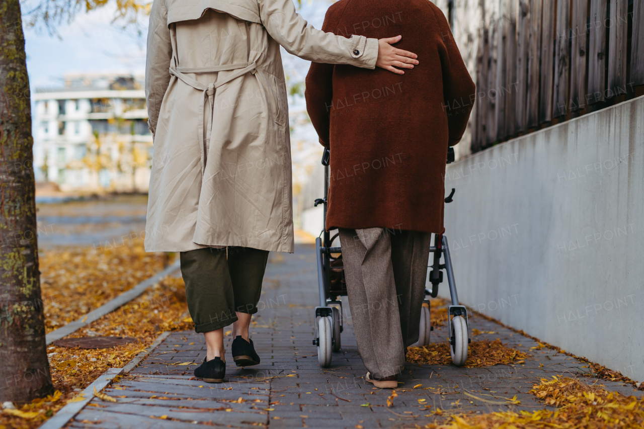 Rear view of grandmother and mature granddaughter on a walk in the city park, during a windy autumn day. Caregiver and senior lady with mobility walker enjoying the fall weather.