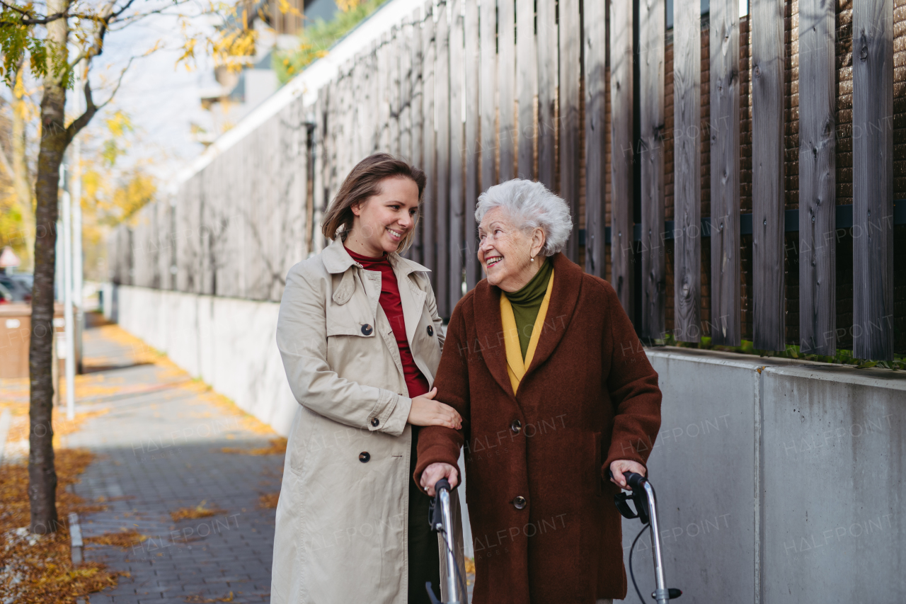 Front view of grandmother and mature granddaughter on a walk in the city park, during a windy autumn day. Caregiver and senior lady with mobility walker enjoying the fall weather.