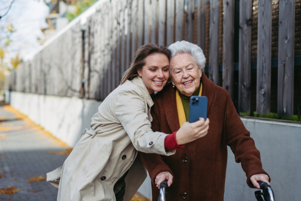 Grandmother and mature granddaughter on walk in city streets, taking selfie. Caregiver and senior lady enjoying the windy autumn weather.