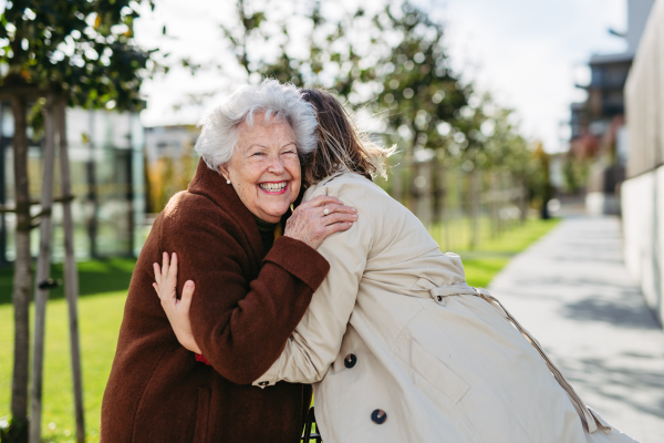 Grandmother and mature granddaughter on a walk in the city park, during a windy autumn day. Caregiver and senior lady enjoying the fall weather, hugging each other.