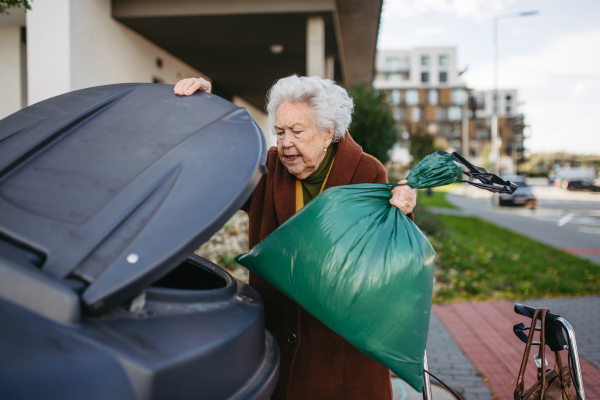 Elderly woman disposing of trash into the garbage can of waste container in front of her apartment complex.