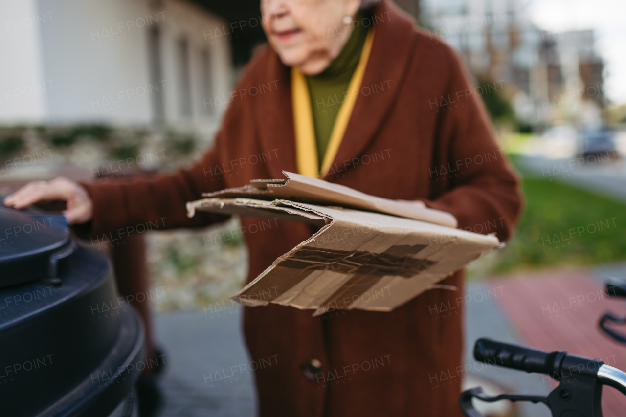 Elderly woman disposing cardboard into the garbage can of waste container in front of her apartment complex.