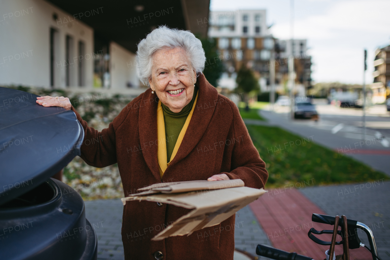 Elderly woman disposing cardboard into the garbage can of waste container in front of her apartment complex.