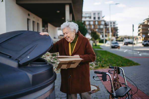 Elderly woman disposing cardboard into the garbage can of waste container in front of her apartment complex.