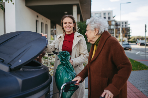 Woman helping elderly neighbor throw away trash into the garbage can, waste container in front of their apartment complex.