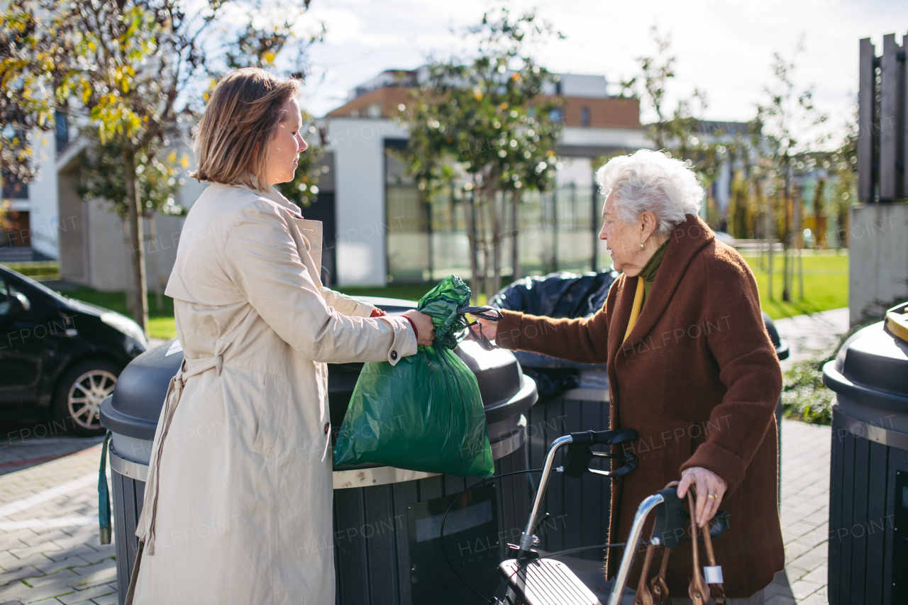 Woman helping elderly neighbor throw away trash into the garbage can, waste container in front of their apartment complex.