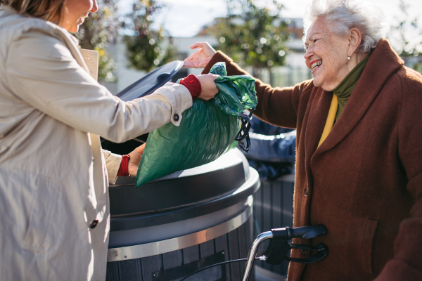 Woman helping elderly neighbor throw away trash into the garbage can, waste container in front of their apartment complex.