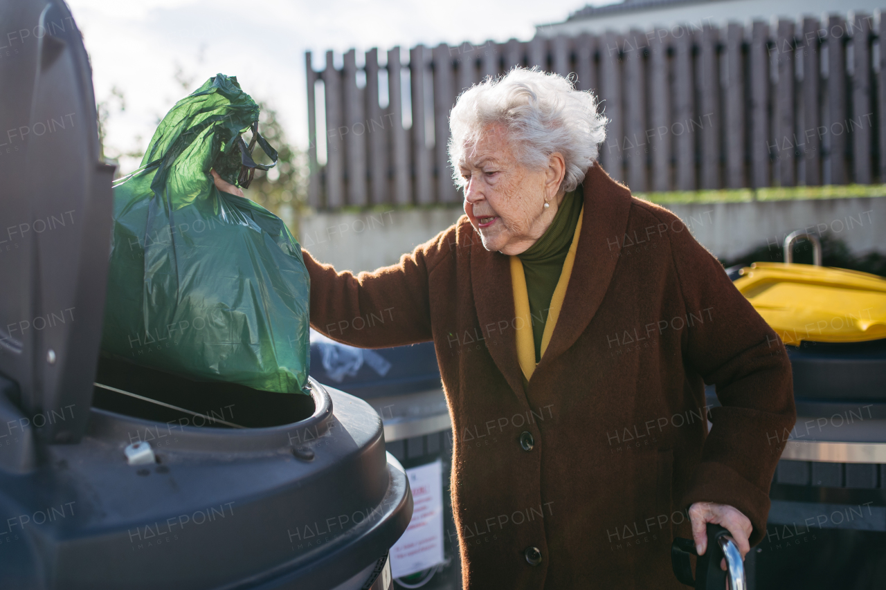 Elderly woman disposing of trash into the garbage can of waste container in front of her apartment complex.