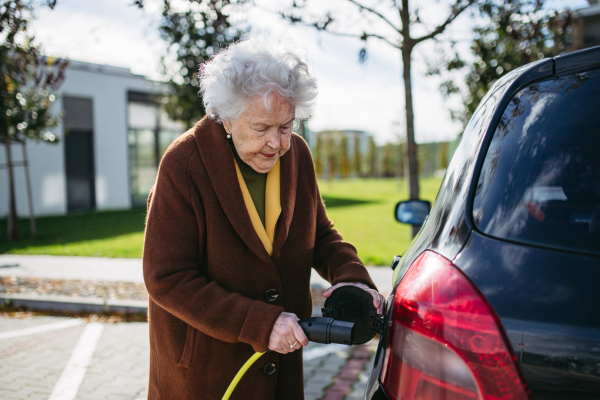 Close up of beautiful senior woman plugging charger in her electric car. Progressive elderly woman charging her electric vehicle on the street.