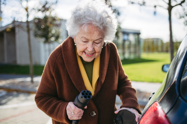 Close up of beautiful senior woman plugging charger in her electric car. Progressive elderly woman charging her electric vehicle on the street.