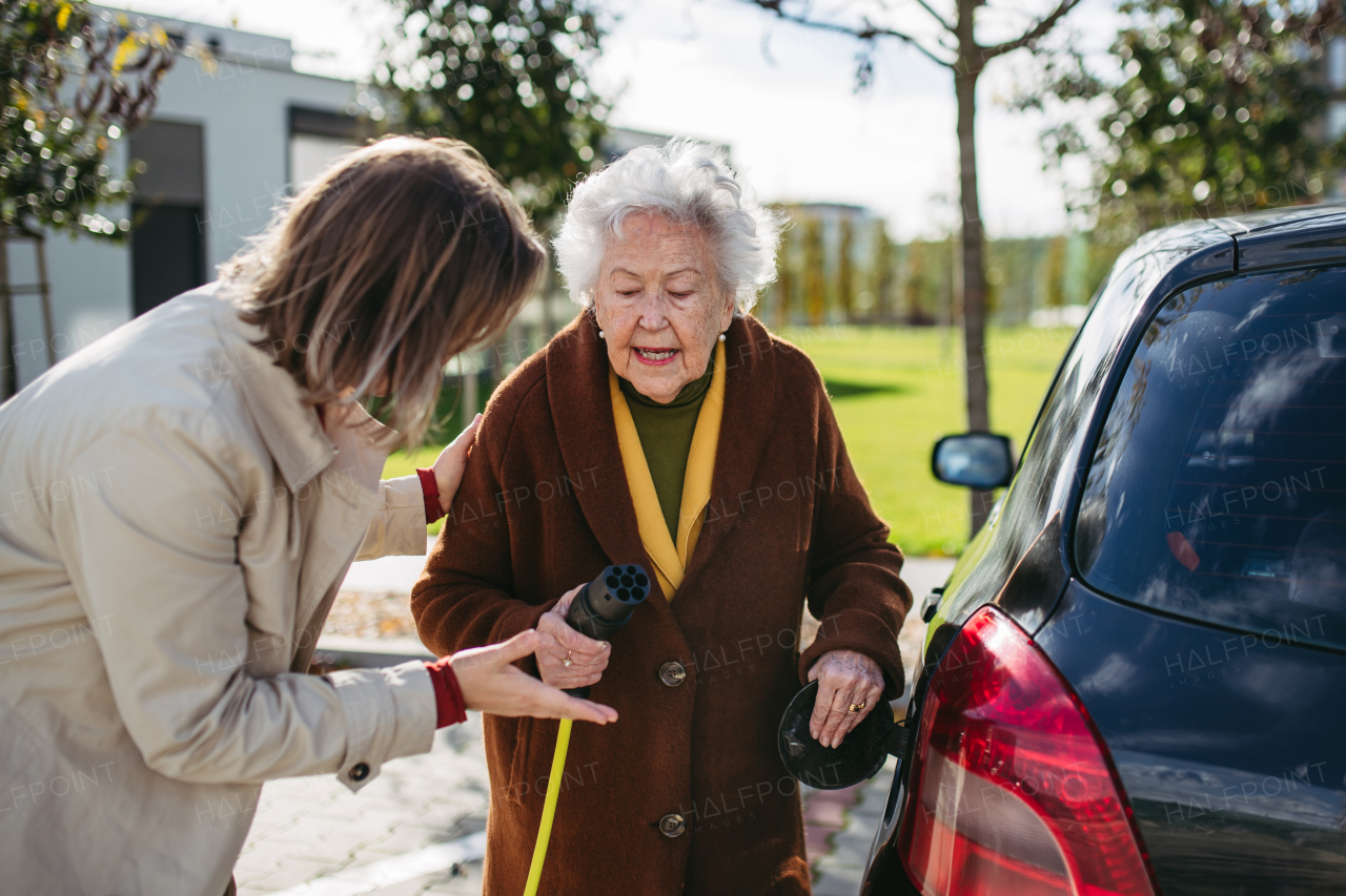 Granddaughter teaching senior woman how to charge new electric car. Progressive grandmother charging her electric vehicle on the street.