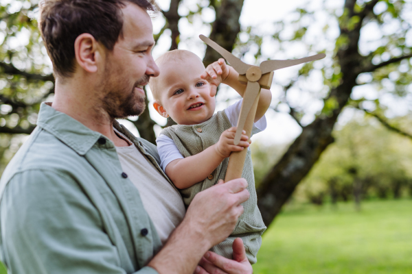 Baby and father holding wind turbine model, outdoors in park. Concept of renewable wind energy, sustainable future for next generation.