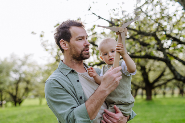 Baby and father holding wind turbine model, outdoors in park. Concept of renewable wind energy, sustainable future for next generation.