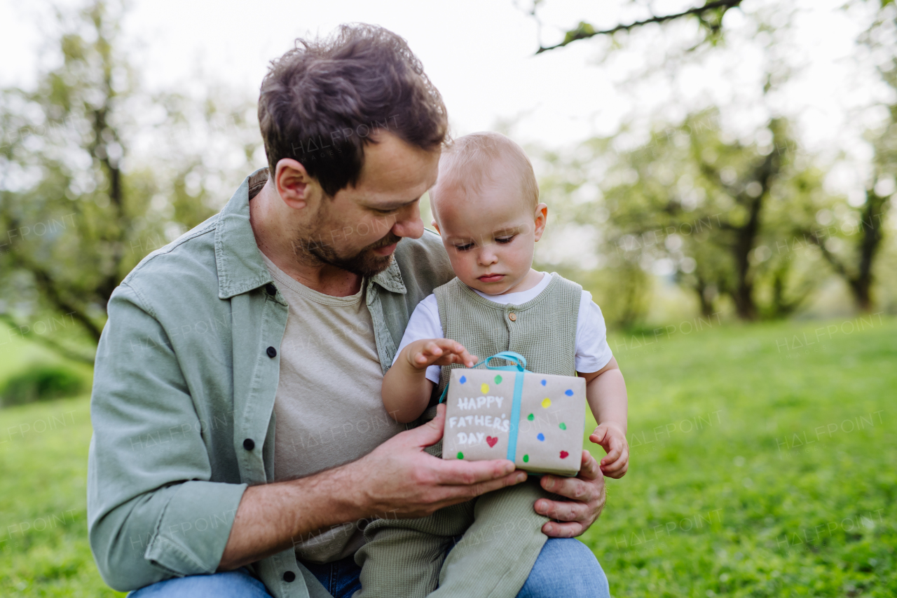 A father holding little baby and gift for Father's day. Family time outdoors in the park during warm spring day.