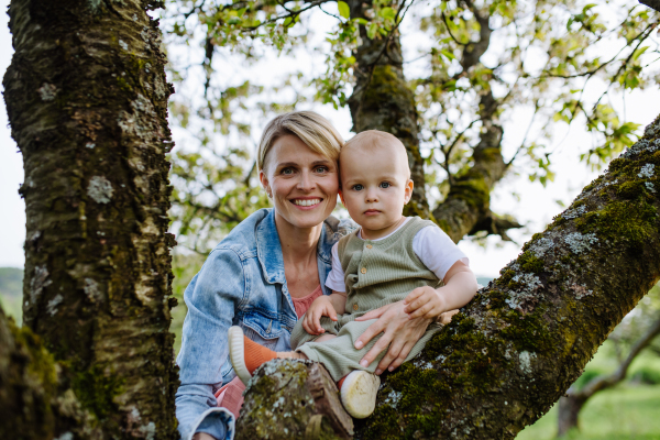 Portrait of beautiful mature first time mother with small toddler, baby, outdoors in spring nature, sitting on tree branch.