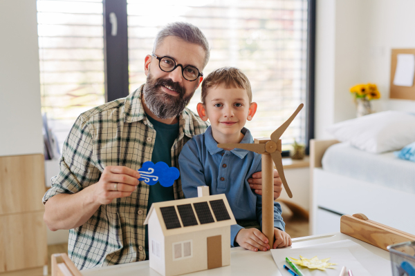 Father explaining renewable green energy, teaching about sustainable lifestyle his young son. Playing with model of house with solar panels, wind turibine at home. Learning through play.