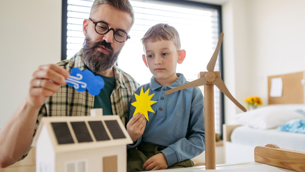 Father explaining renewable green energy, teaching about sustainable lifestyle his young son. Playing with model of house with solar panels, wind turibine at home. Learning through play.