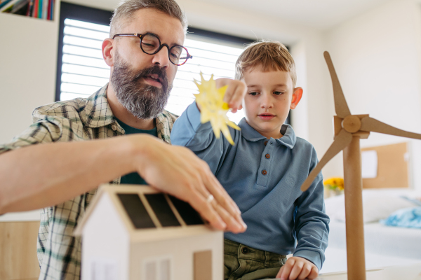 Father explaining renewable green energy, teaching about sustainable lifestyle his young son. Playing with model of house with solar panels, wind turibine at home. Learning through play.