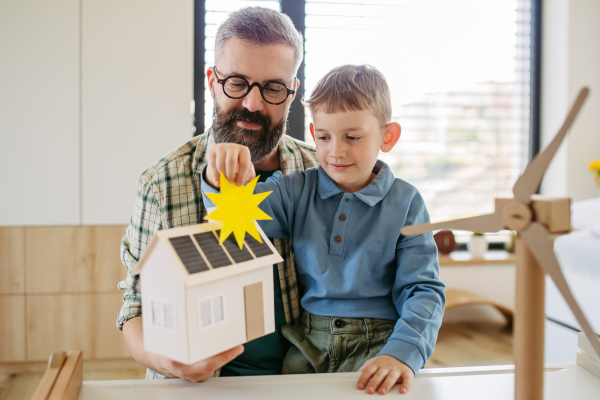 Father explaining renewable green energy, teaching about sustainable lifestyle his young son. Playing with model of house with solar panels, wind turibine at home. Learning through play.