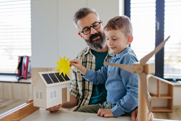 Father explaining renewable green energy, teaching about sustainable lifestyle his young son. Playing with model of house with solar panels, wind turibine at home. Learning through play.