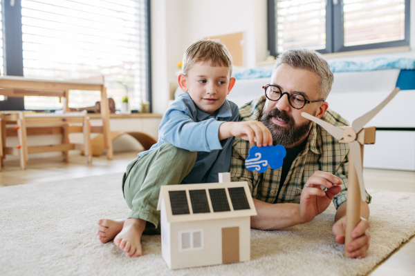 Father explaining renewable green energy, teaching about sustainable lifestyle his young son. Playing with model of house with solar panels, wind turibine at home. Learning through play.