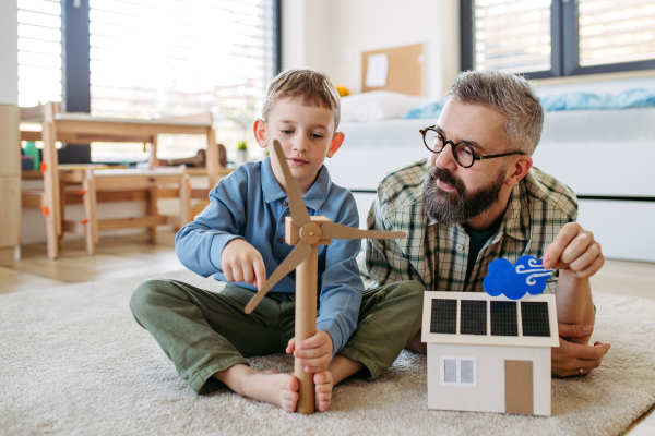 Father explaining renewable green energy, teaching about sustainable lifestyle his young son. Playing with model of house with solar panels, wind turibine at home. Learning through play.