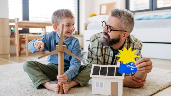 Father explaining renewable green energy, teaching about sustainable lifestyle his young son. Playing with model of house with solar panels, wind turibine at home. Learning through play.