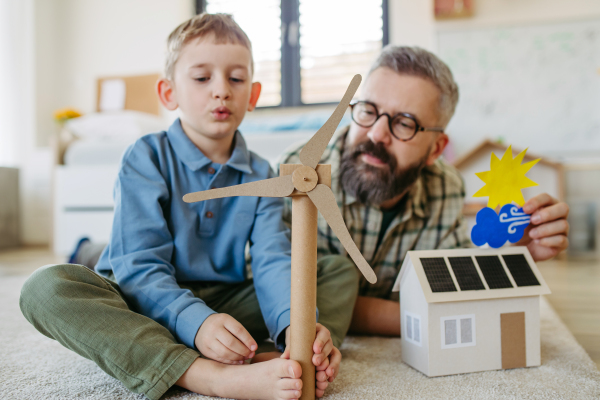 Father explaining renewable green energy, teaching about sustainable lifestyle his young son. Playing with model of house with solar panels, wind turibine at home. Learning through play.