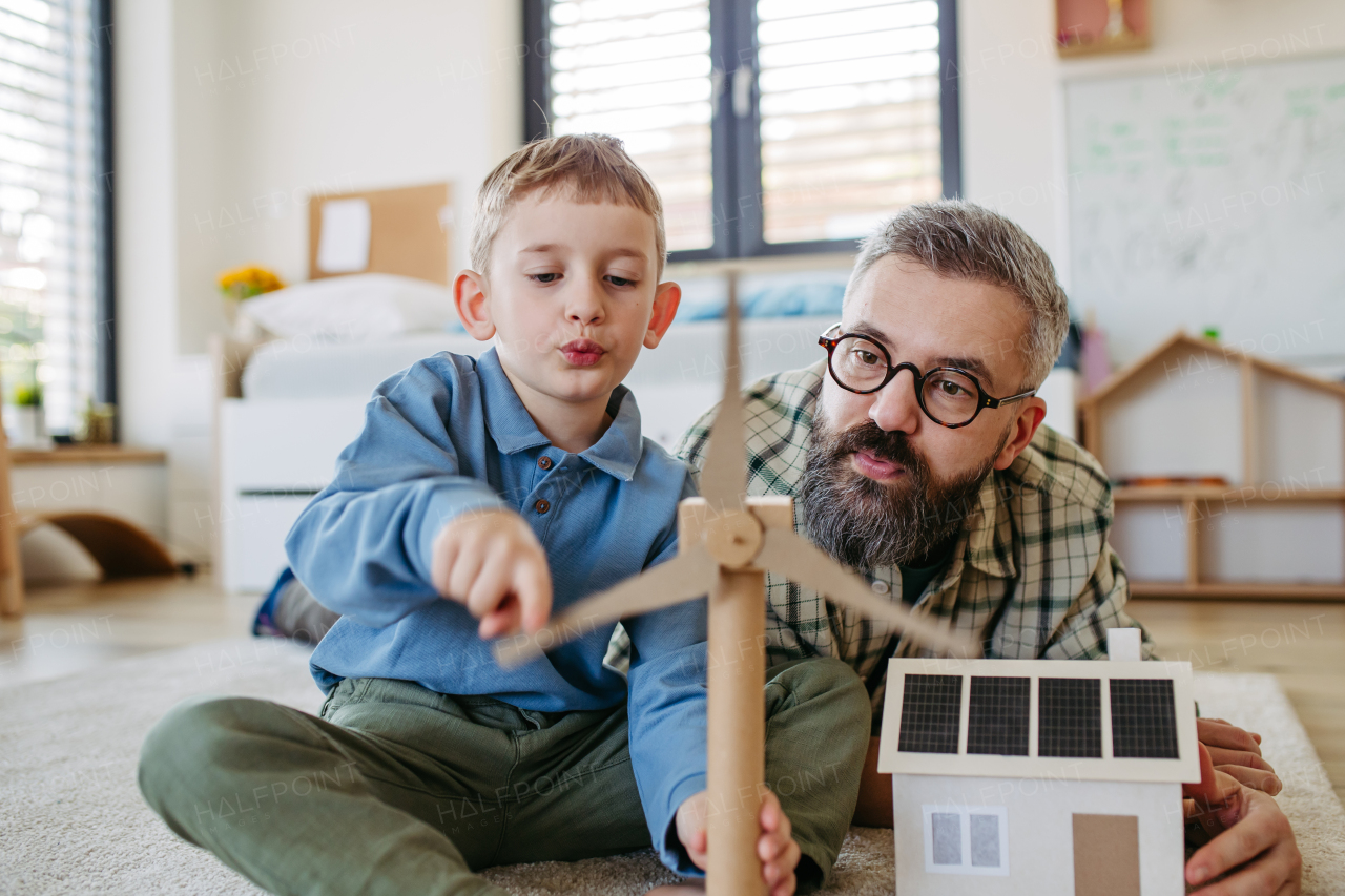 Father explaining renewable green energy, teaching about sustainable lifestyle his young son. Playing with model of house with solar panels, wind turibine at home. Learning through play.