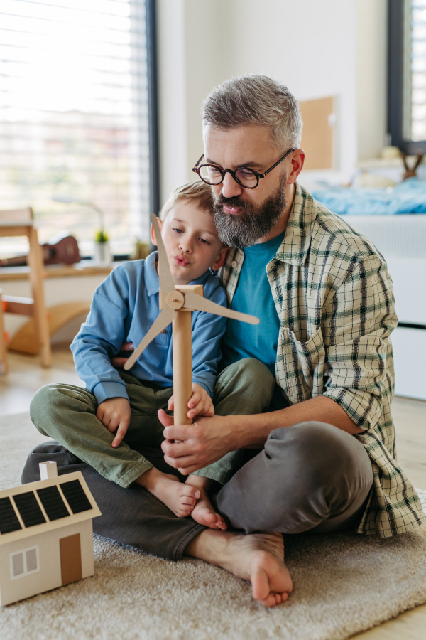 Father explaining renewable green energy, teaching about sustainable lifestyle his young son. Playing with model of house with solar panels, wind turibine at home. Learning through play.