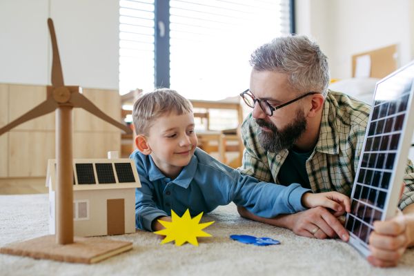 Father explaining renewable green energy, teaching about sustainable lifestyle his young son. Playing with model of house with solar panels, wind turibine at home. Learning through play.