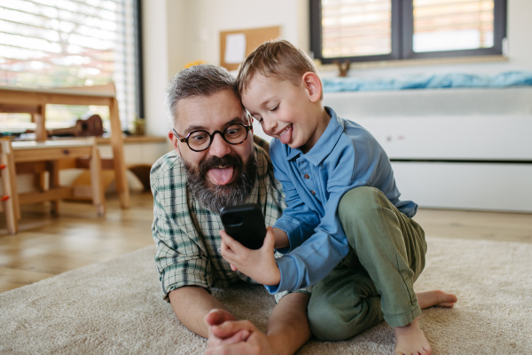 Little boy making selfie on smartphone with father, lying on the floor in kids room, making silly faces. Dad explaining technology to son, digital literacy for kids.