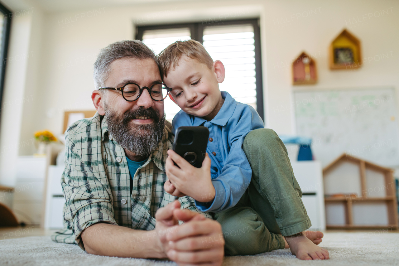 Little boy watching cartoon movie on smartphone with father, lying on floor in kids room. Dad explaining technology to son, digital literacy for a kids.