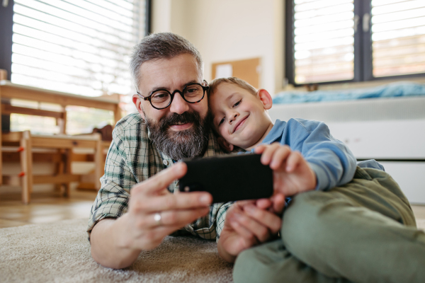 Little boy watching cartoon movie on smartphone with father, lying on floor in kids room. Dad explaining technology to son, digital literacy for a kids.