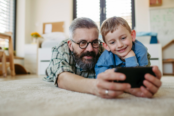 Little boy making selfie on smartphone with father, lying on the floor in kids room, making silly faces. Dad explaining technology to son, digital literacy for kids.