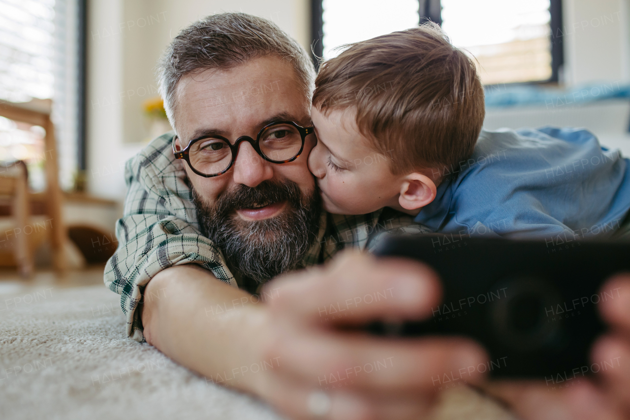 Little boy making selfie on smartphone with father, lying on the floor in kids room, making silly faces. Dad explaining technology to son, digital literacy for kids.