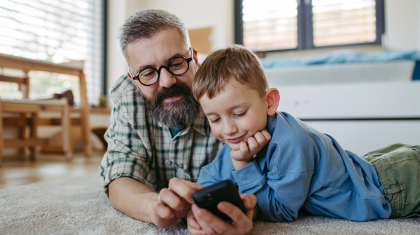 Little boy watching cartoon movie on smartphone with father, lying on floor in kids room. Dad explaining technology to son, digital literacy for a kids.