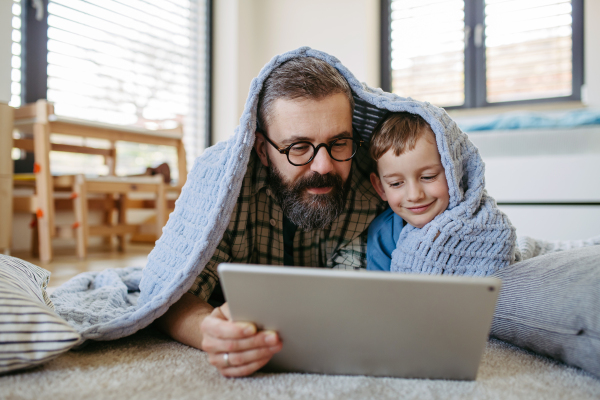 Little boy watching cartoon movie on the tablet with father, lying under blanket on floor in kids room. Dad explaining technology to son, digital literacy for kids.