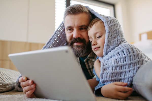 Little boy watching cartoon movie on the tablet with father, lying under blanket on floor in kids room. Dad explaining technology to son, digital literacy for kids.