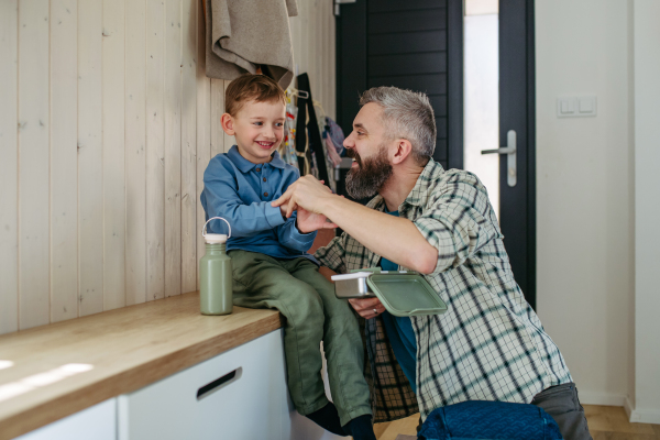 Father helping son get ready for kindergarten, preschool. Putting coat and shoes on, handing lunchbox with snacks.