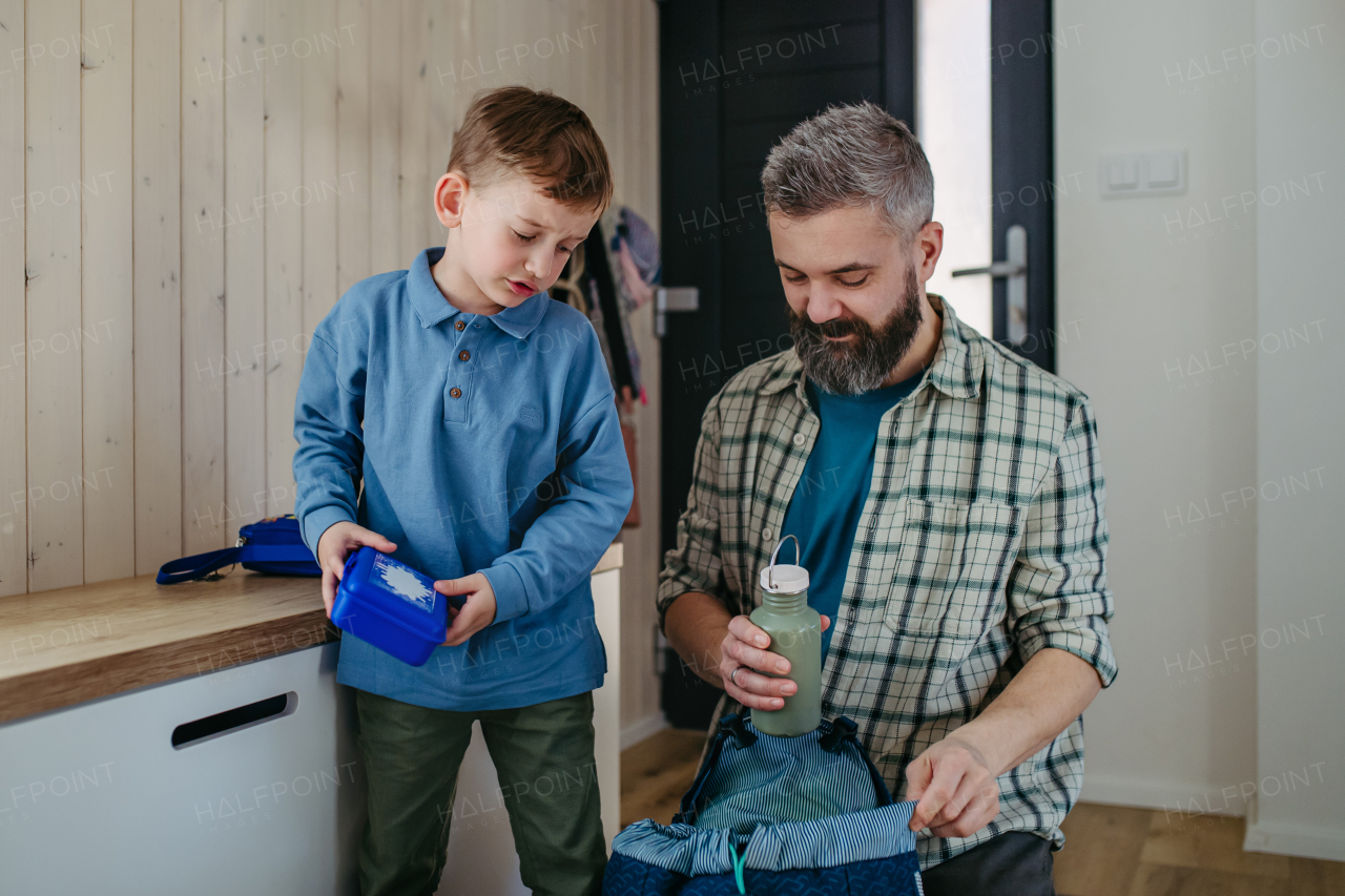 Father helping son get ready for kindergarten, preschool. Putting lunchbox with food, snacks in his backpack.