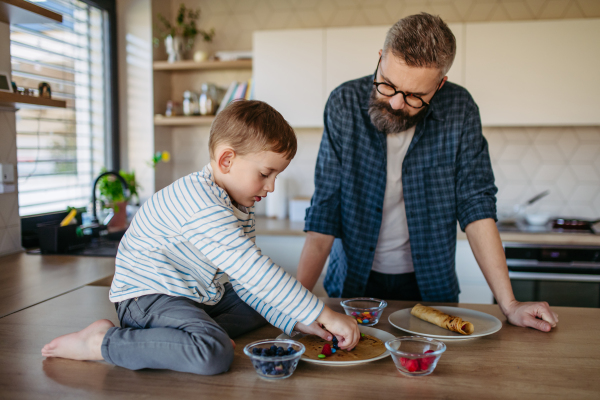 Boy filling pancake with fruit, sweets. Father spending time with son at home, making snack together, cooking. Weekend activities for single dad with young boy. Fathers day concept.