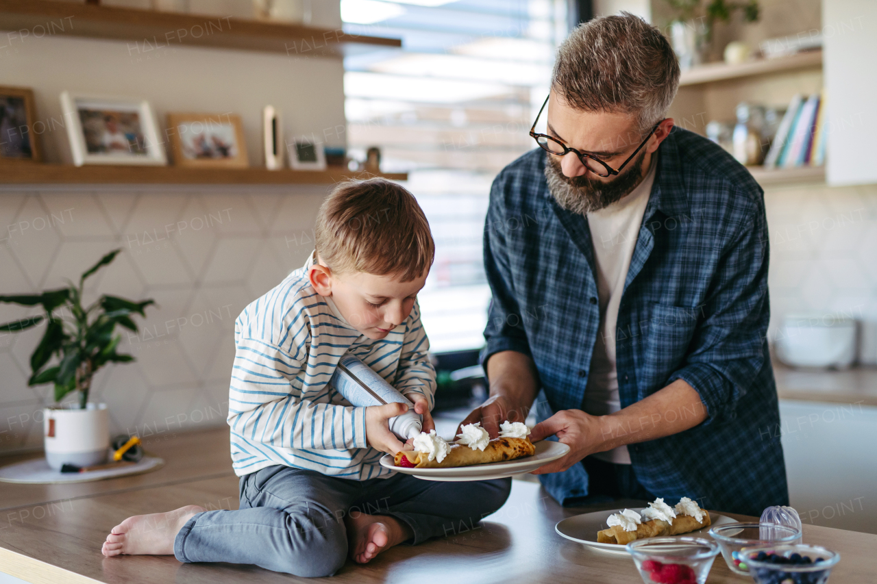 Boy filling pancake with fruit, sweets. Father spending time with son at home, making snack together, cooking. Weekend activities for single dad with young boy. Fathers day concept.