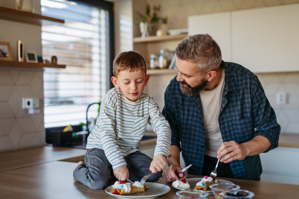 Boy filling pancake with fruit, sweets. Father spending time with son at home, making snack together, cooking. Weekend activities for single dad with young boy. Fathers day concept.