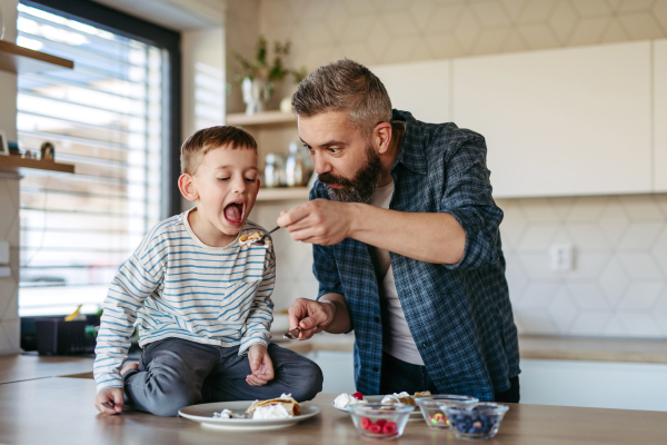 Boy eating pancake with fruit, sweets. Father spending time with son at home, making snack together, cooking. Weekend activities for single dad with young boy. Fathers day concept.