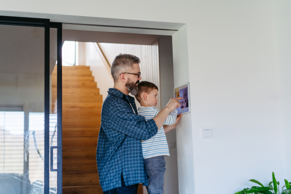 Father showing son smart home system, adjusting household functions, lighting, security cameras, door locks and thermostat or heating settings.