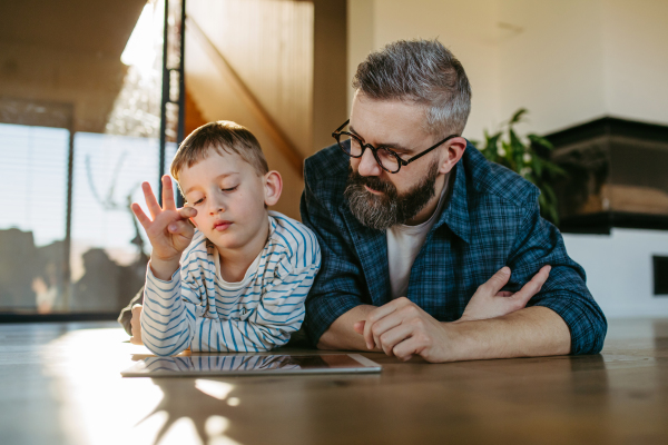 Little boy watching cartoon movie on the tablet with father, lying uon the floor. Dad explaining technology to son, digital literacy for kids.