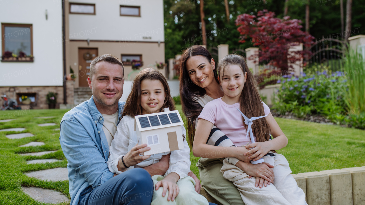 Happy family holding model of house with solar panels on roof and wind turbine model. Alternative, renewable, green energy and sustainable lifestyle concept.