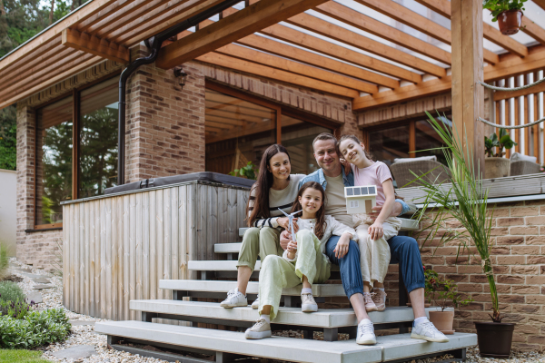 Happy family holding model of house with solar panels on roof and wind turbine model. Alternative, renewable, green energy and sustainable lifestyle concept.