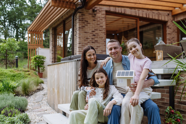 Happy family holding model of house with solar panels on roof and wind turbine model. Alternative, renewable, green energy and sustainable lifestyle concept.